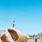 Man on top of large red boulder with blue skies and man on ground