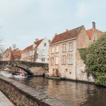 small white boat in canal driving under cobblestone bridge by an old brick building
