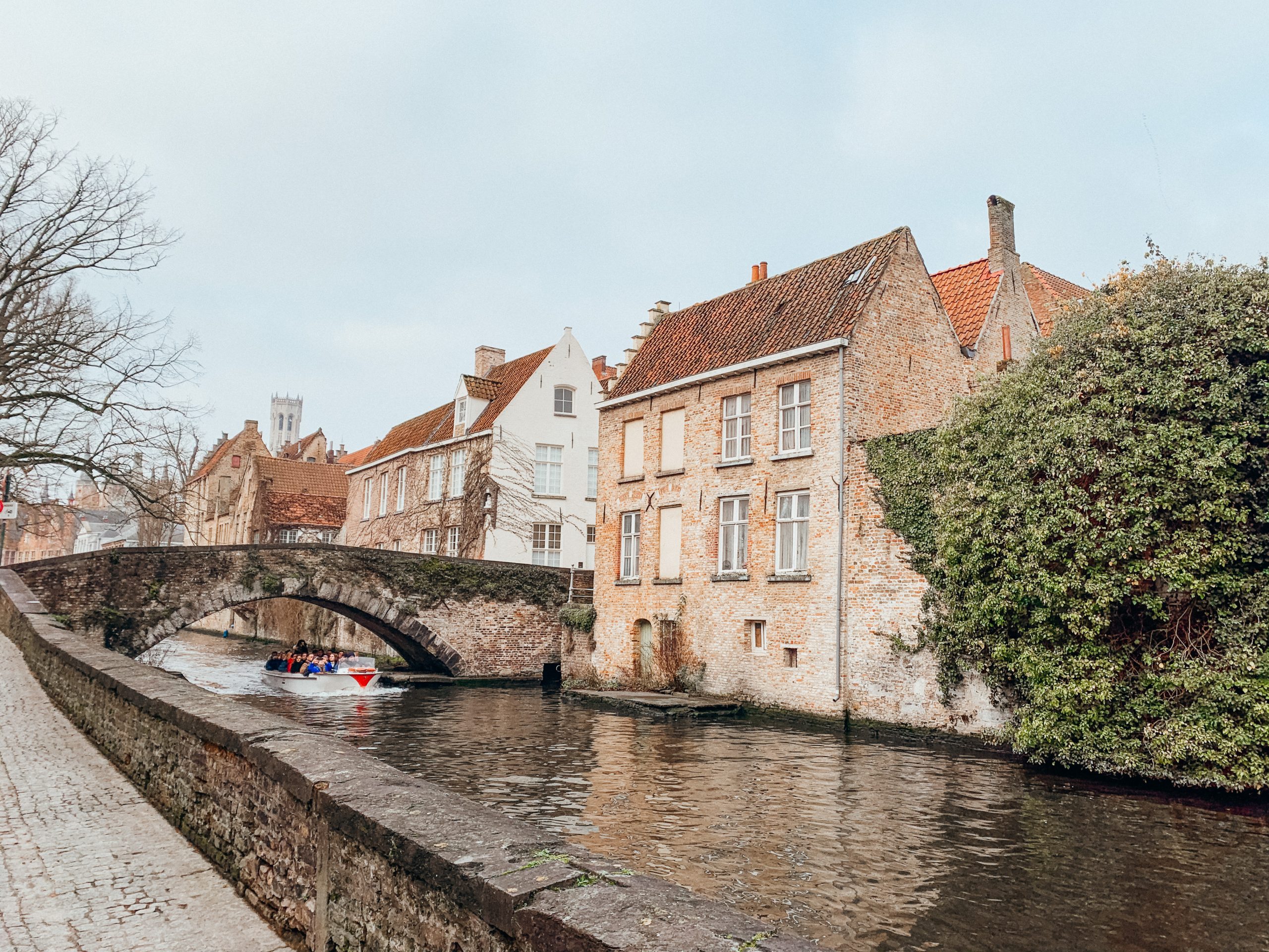 small white boat in canal driving under cobblestone bridge by an old brick building