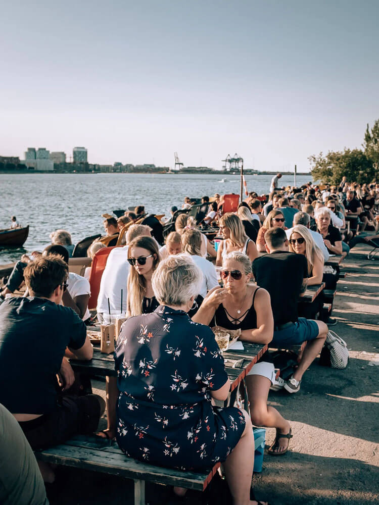 People sit at wooden picnic tables along a harbour in Copenhagen Denmark