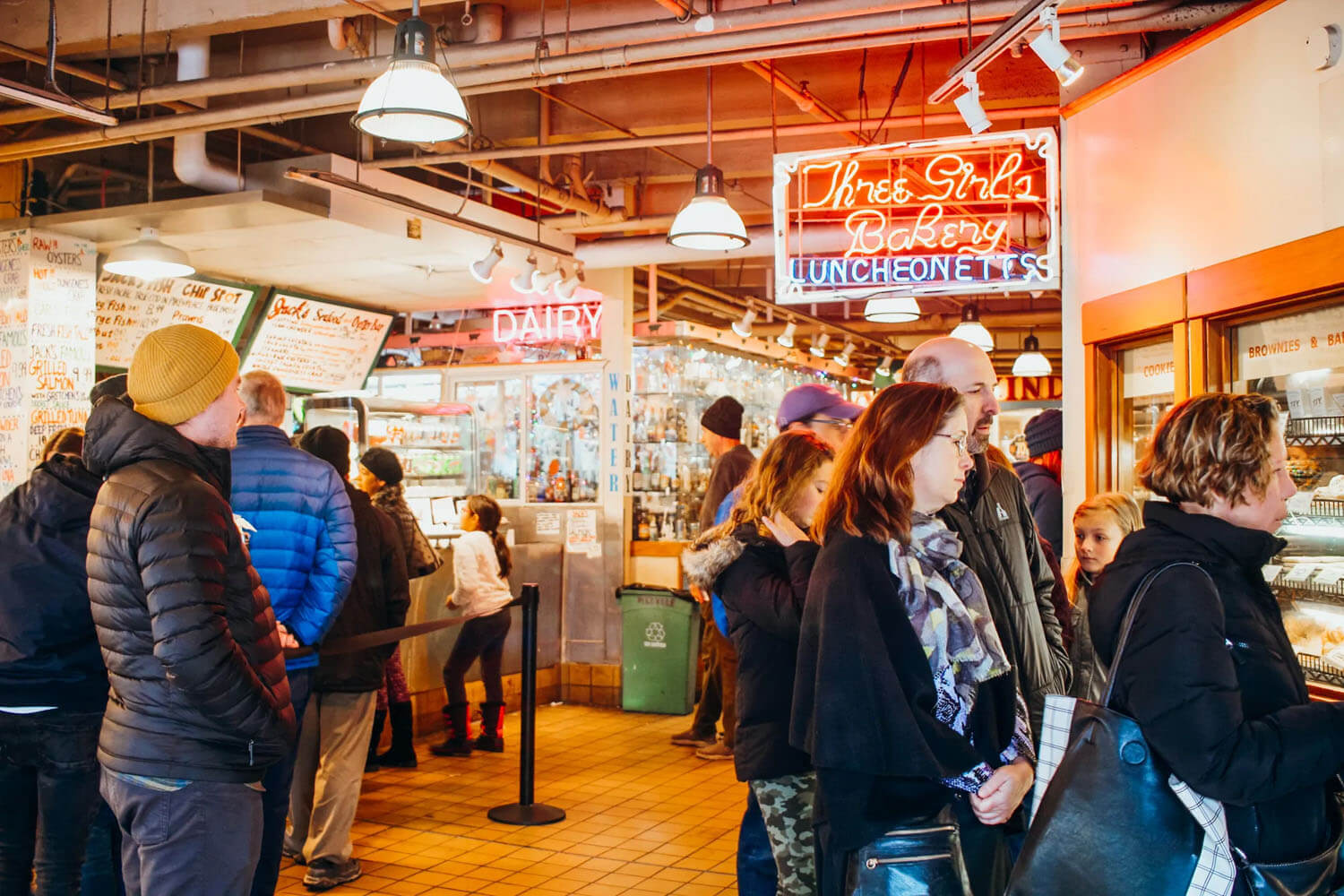 People stand under neon red sign saying 'Three Girls Bakery" looking into bakery window