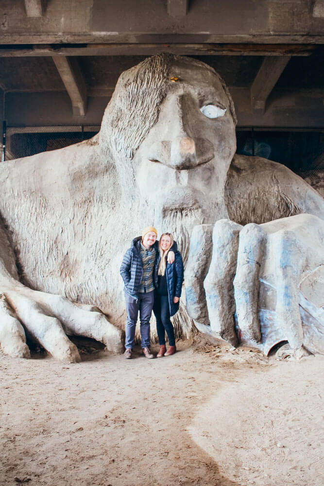 a man and woman stand in front of large sculpture of a troll under an overpass