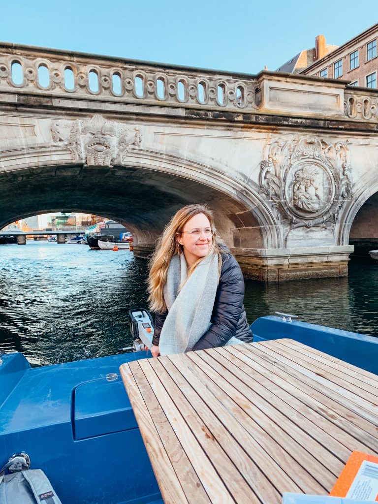 Woman sits on a small boat next to a wooden table going under a bridge