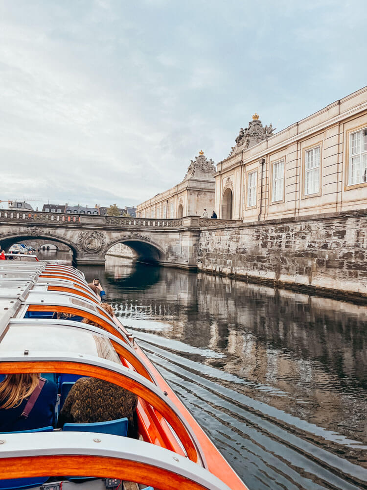 Long orange and white boat in canal about to go under an ornate bridge next to a white ornate building