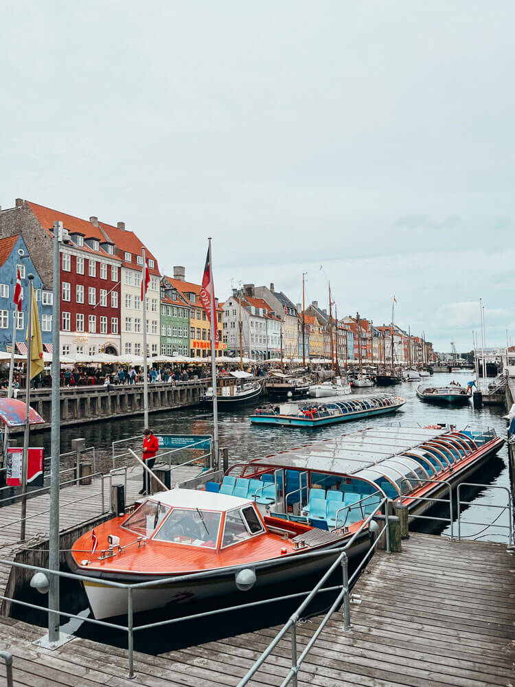 Long, low orange and white boate with blue seats docked in a canal lined with tall colorful buildings