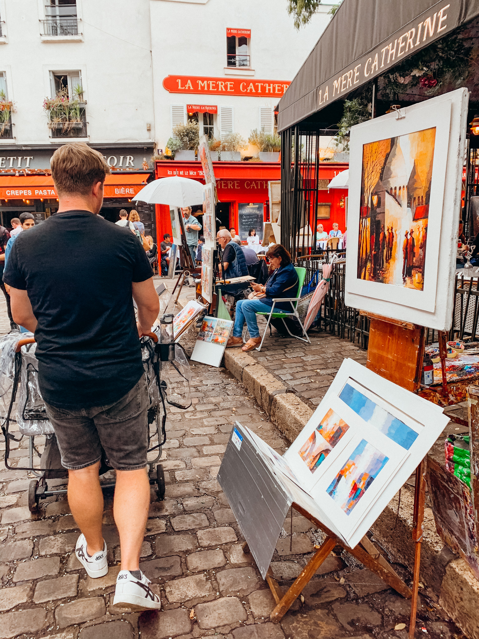 Man pushing stroller past paintings for sale by street vendors