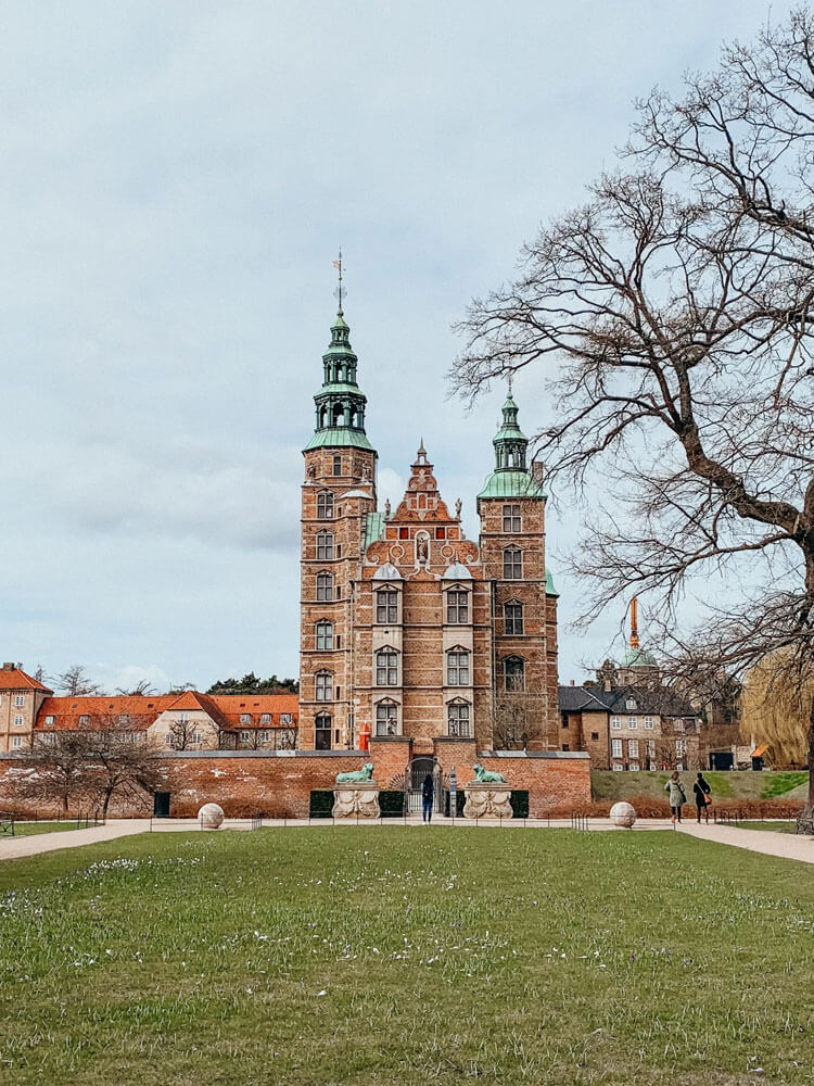 Brown castle with green spires surrounded by grass and orange-roofed buildings
