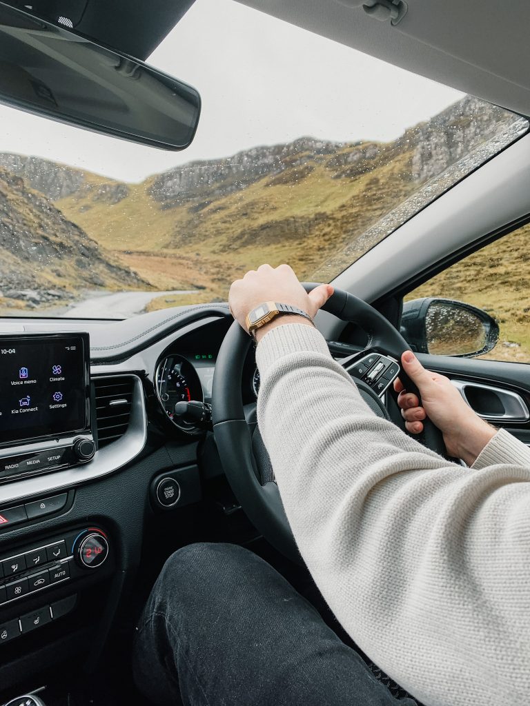 Two hands on a steering wheel with a view of a green and black cliff through the windshield
