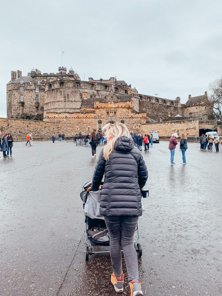 Woman in black coat and pants pushing a grey stroller towards a castle