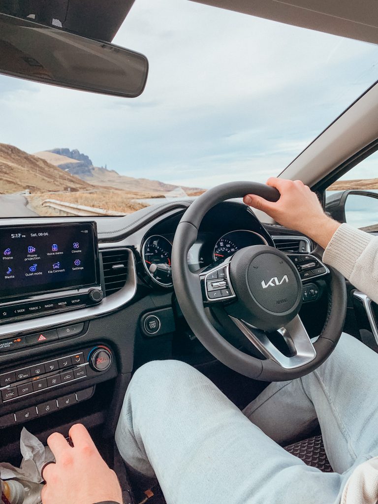 Person with blue pants and white sweater with hands on a black steering wheel driving in the mountains