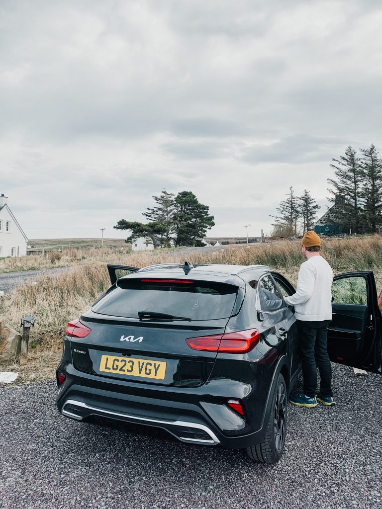Man in white sweater and yellow hat gets into black car parked on small rocks next to a field of grass