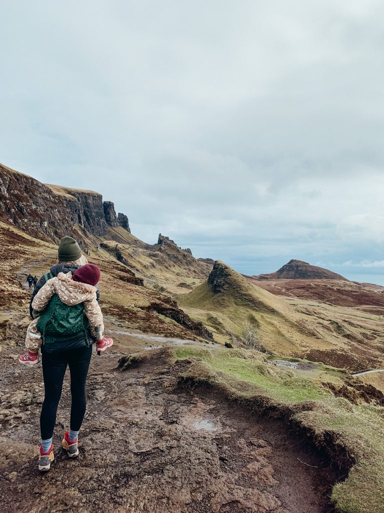 Woman with a toddler strapped to her back overlooks large green cliffs of the Quiraing