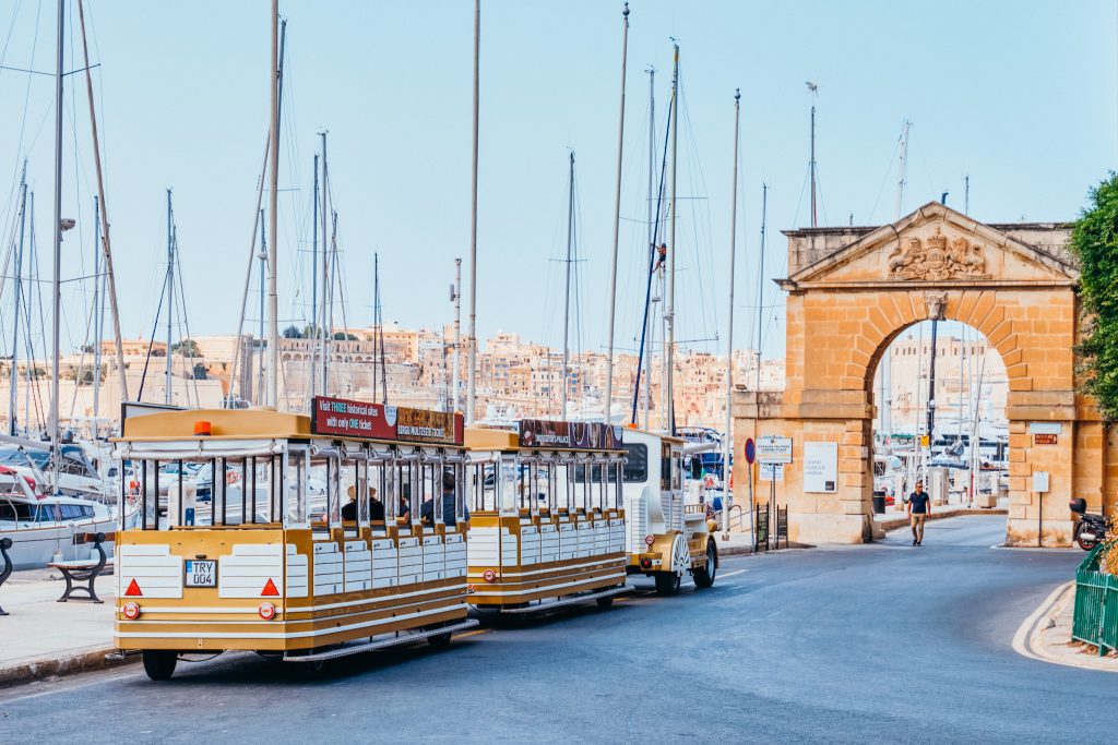 Small white and gold train on wheels sits on the street in front of a stone arch next to a harbor