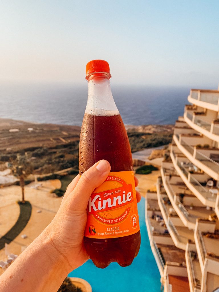 Hand holds a orange soda over a view of a swimming pool and sea
