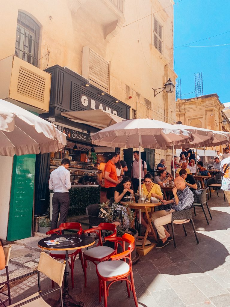 Round tables with umbrellas in front of a stone building with a black storefront with a sign that reads 'Grano'