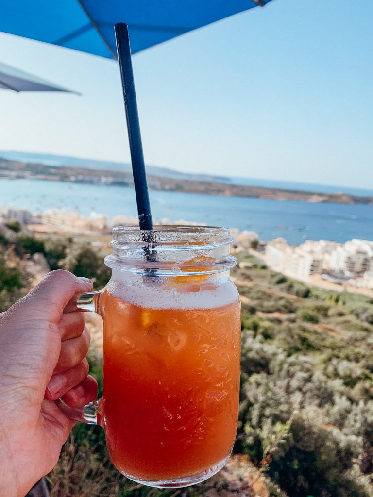 Hand holds a glass mug filled with a red juice with the sea in the background