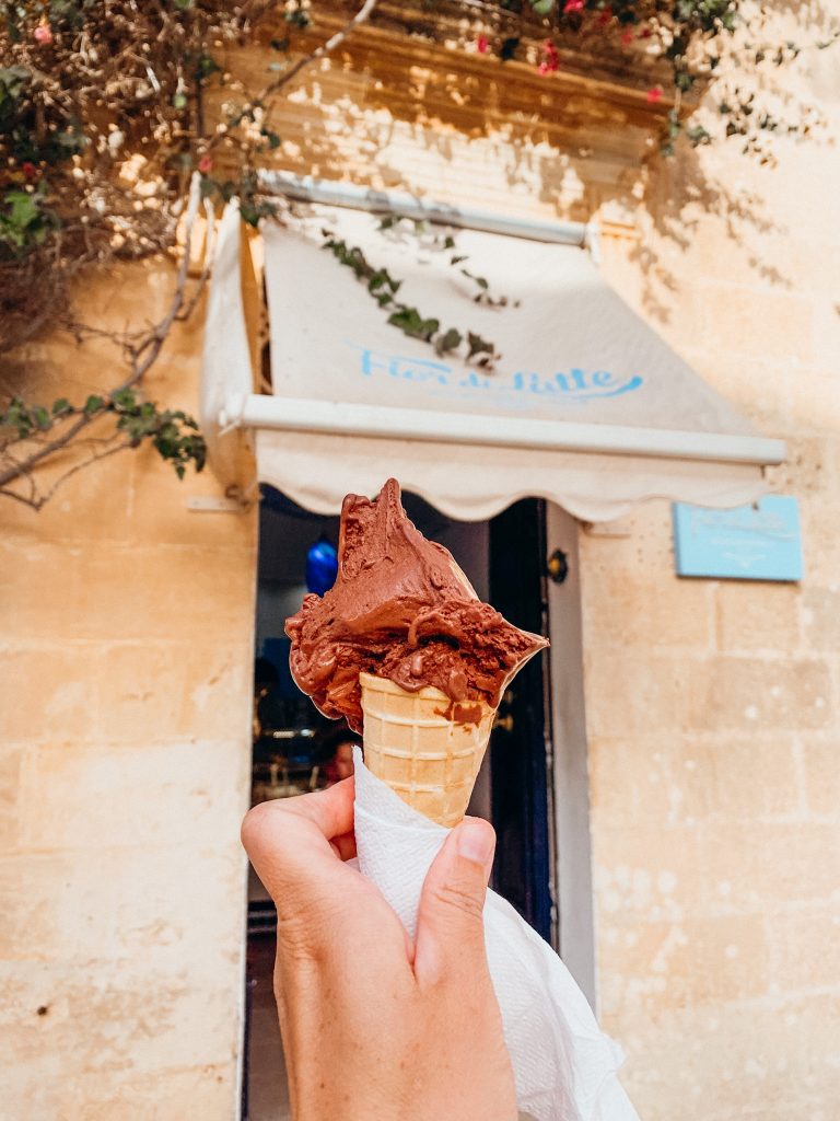 A hand holding an ice cream cone in front of a ice cream store front in a stone building