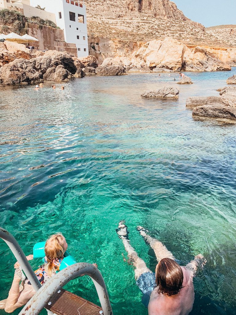 Child and father float in green and blue water with rocks and cliffs in teh distance