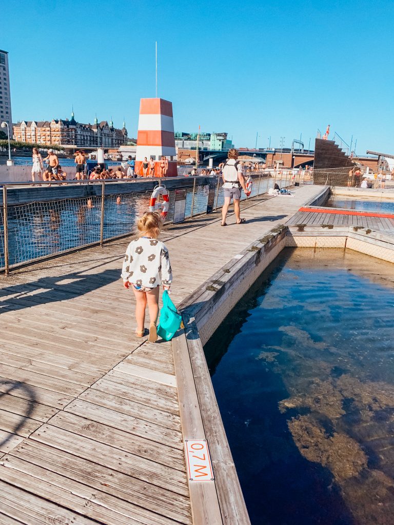 Child and man walk along wooden walkway between two pools of water at a Copenhagen harbor bath