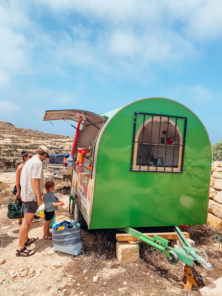 People order smoothies from small green food stand on dirt in Gozo