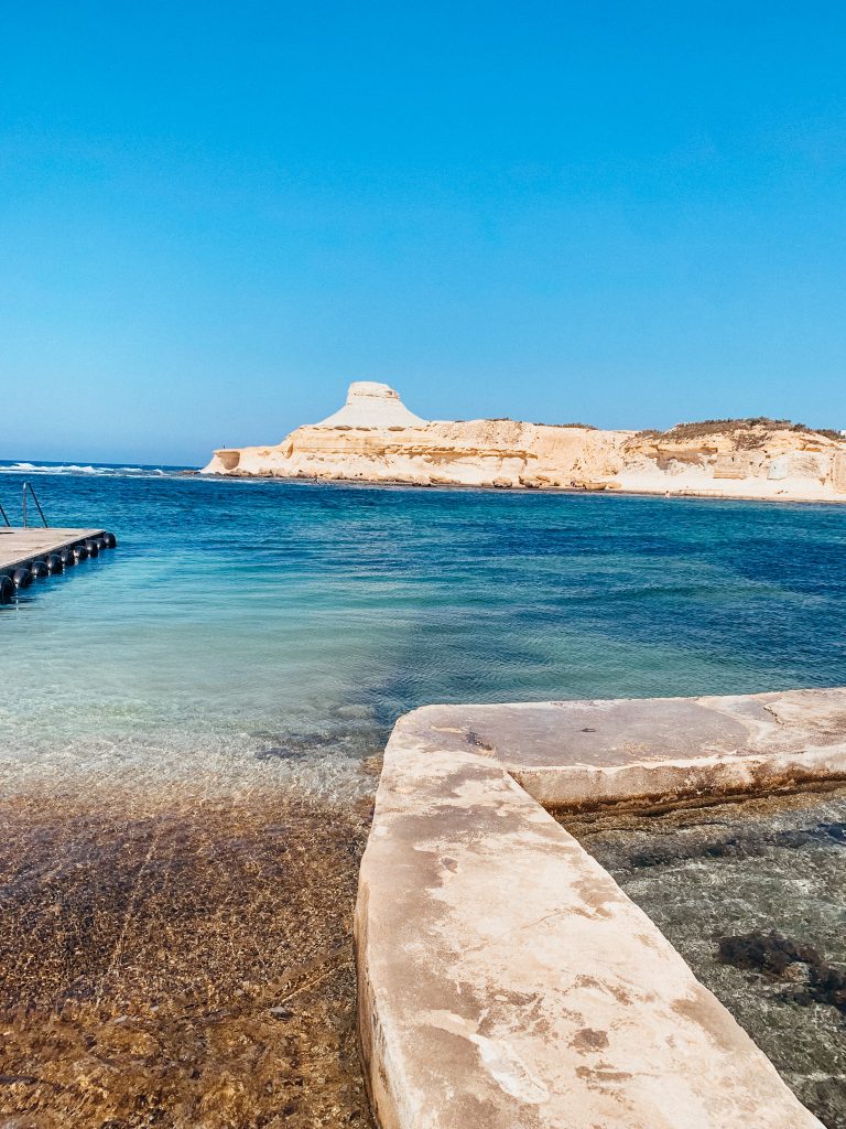 Cement path leads to bright blue sea water with large white, stone hill in the distance