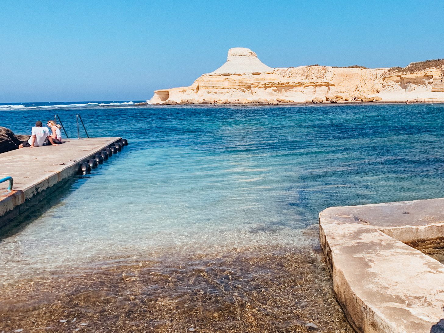 Cement path leads to bright blue water with large white, stone hill in the distance