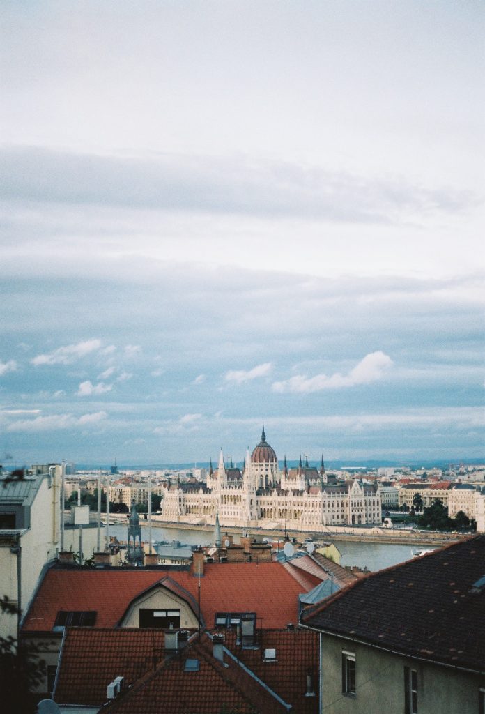 Red shingles on a home in the foreground and a large tan building with multiple spires along a riverbankin the background