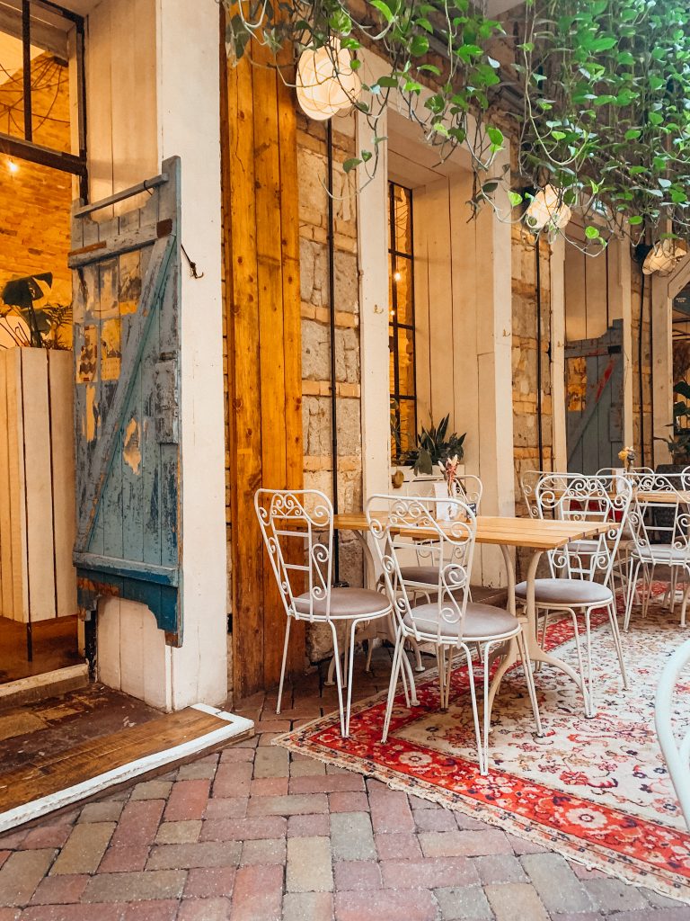 White, curled iron chairs at a brown table on a red and white flowered rug next to a brick and wood wall in a trendy restaurant in Budapest Hungary