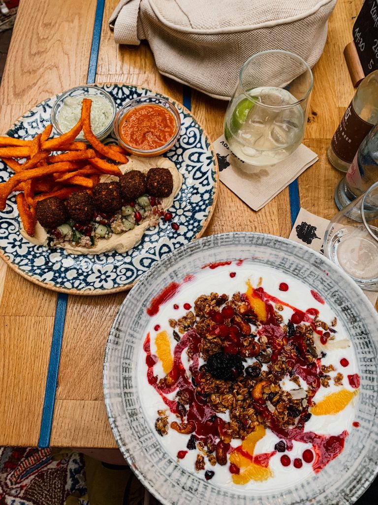 Two plates of food on a wooden table. One has falafel and sweet potato fries while the other has white yogurt with granola and fruit