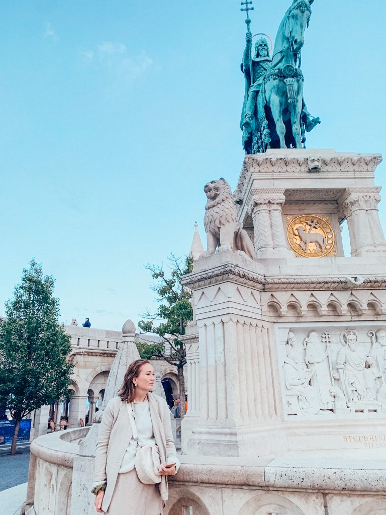 Woman in tan stands in front of tan statue of a lion with a blue statue of a lion above