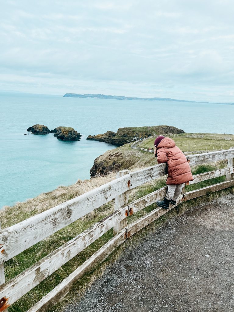 Young girl stands on wooden fence overlooking green cliffs and blue water