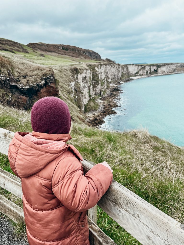 Young child stands next to wooden fence looking at views of white and green cliffs meeting the blue sea