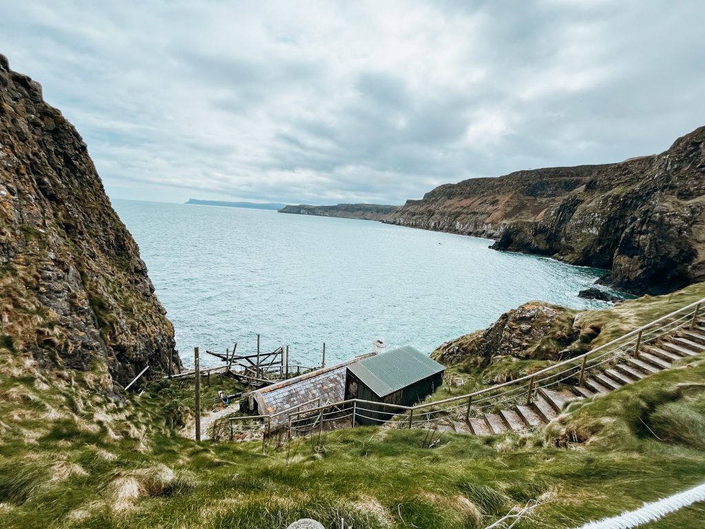 Paths lead down grassy cliffside to old building on the cliffs edge with views of cliffs and the sea in the background
