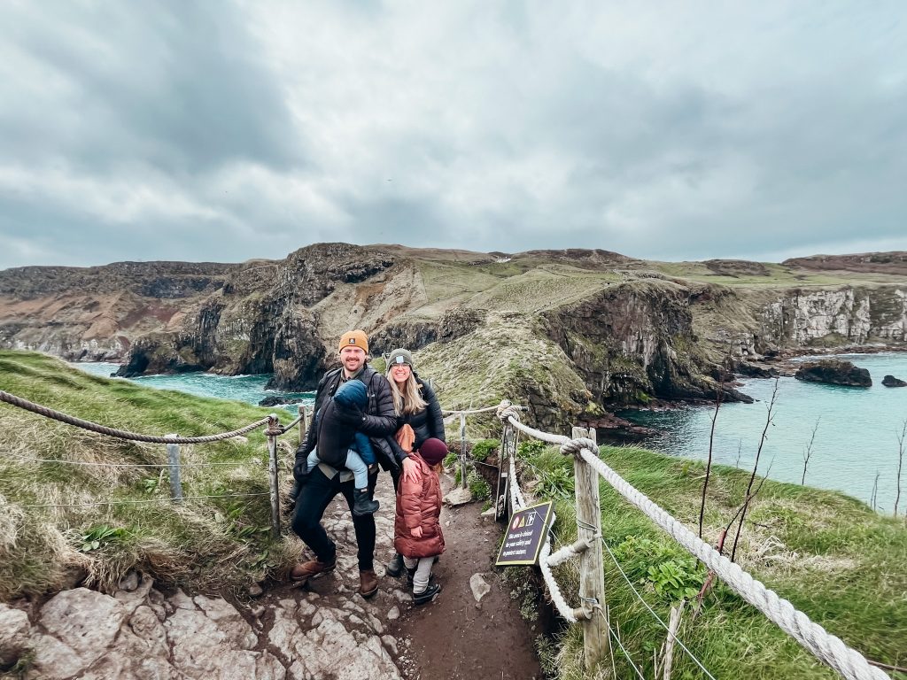 A young family stands on a dirt trail with green and cliffs behind them and views of the sea