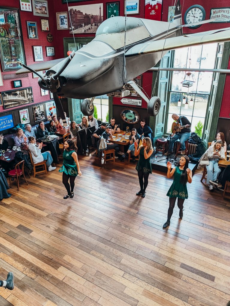Three woman in green dance on a wooden floor surrounded by people sitting at tables