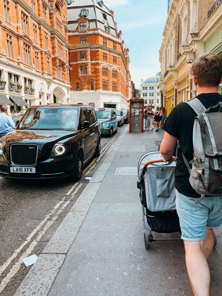 Man pushes a stroller down a sidewalk in London with kids past black cabs and old buildings