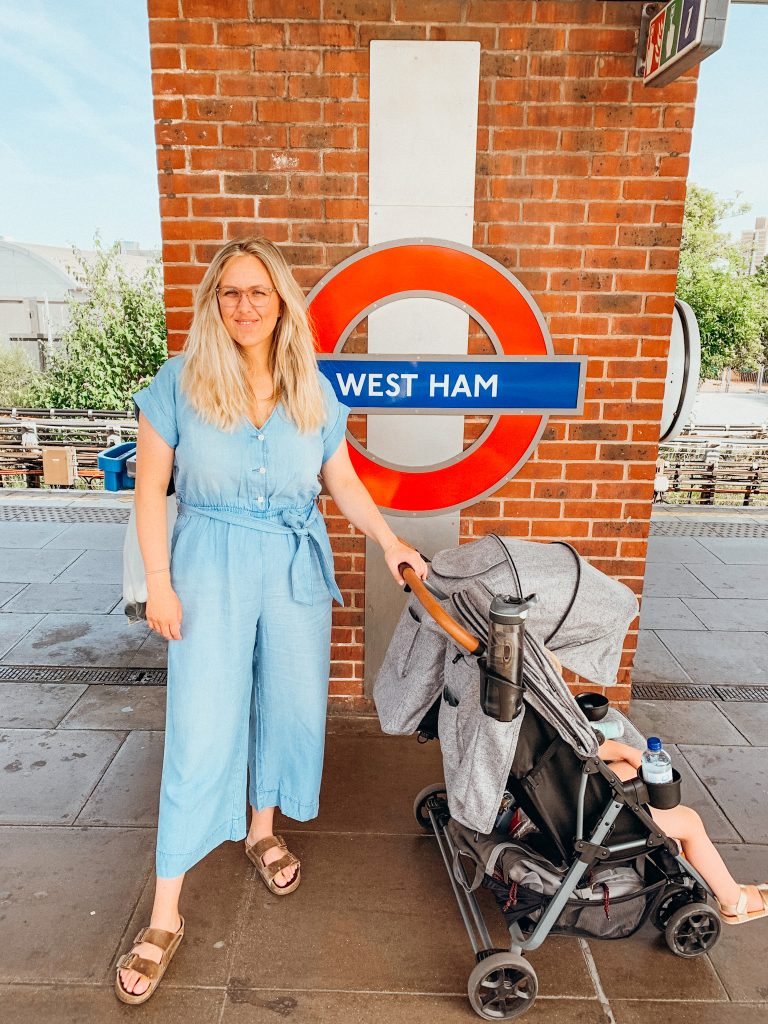 Woman stands with a hand on the stroller in front of sign that says 'West Ham' 