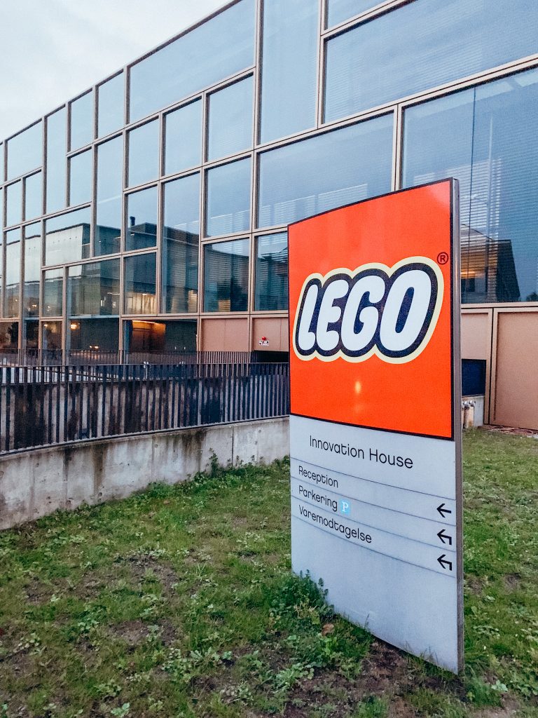 Metal sign with red LEGO logo in front of building full of windows
