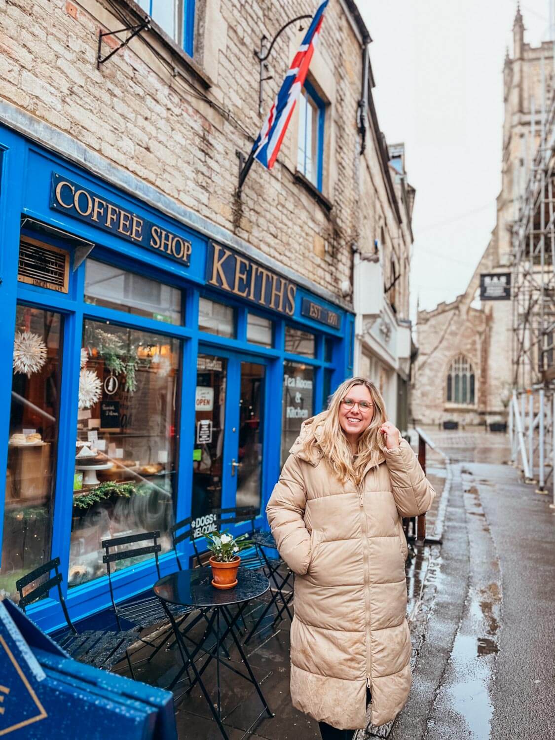 Woman in tan coat stands in front of old brick building with blue window frames and a sign that reads 'Keith's'