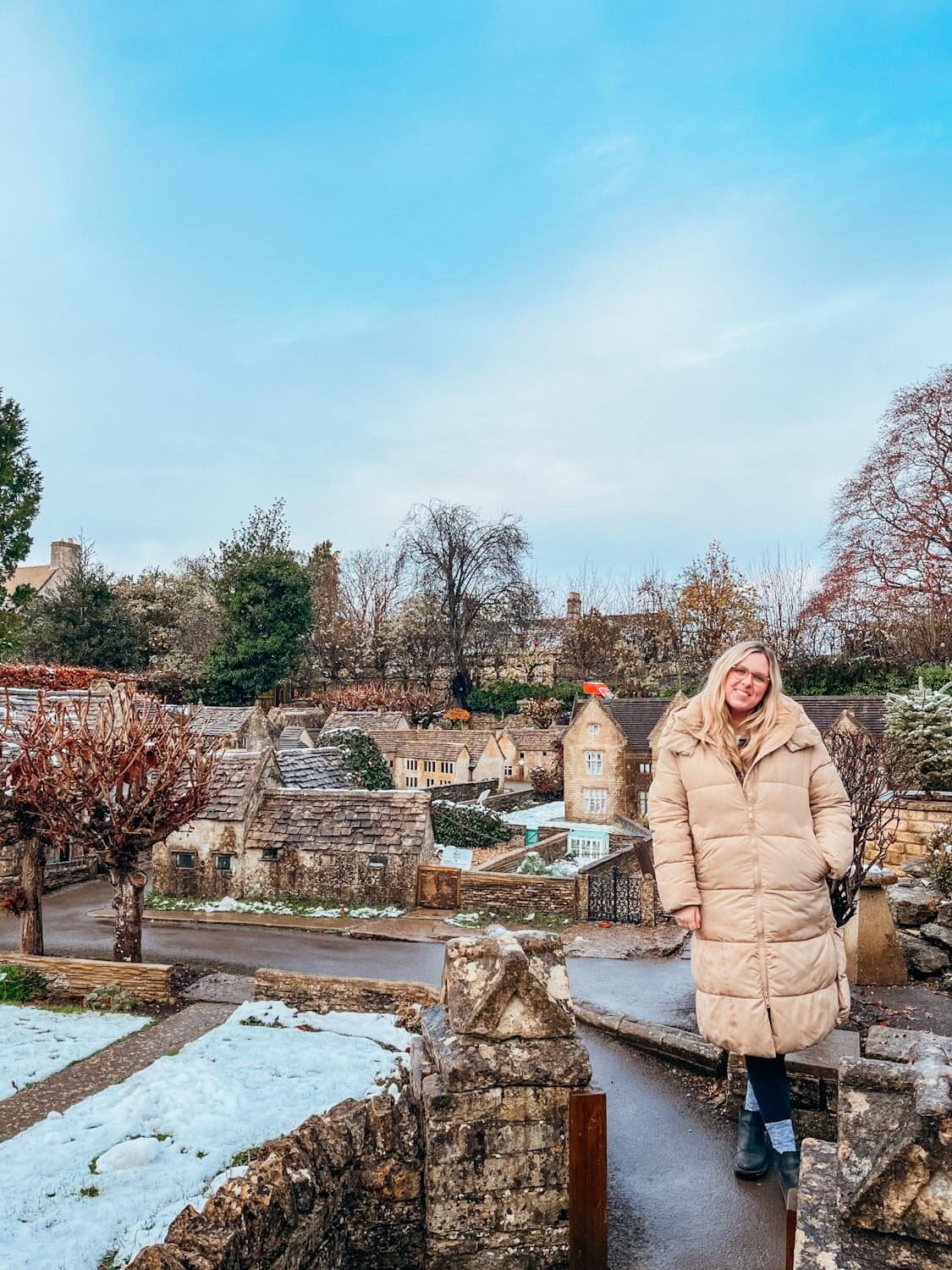 Woman in tan coat stands in front of small replica buildings made of stone