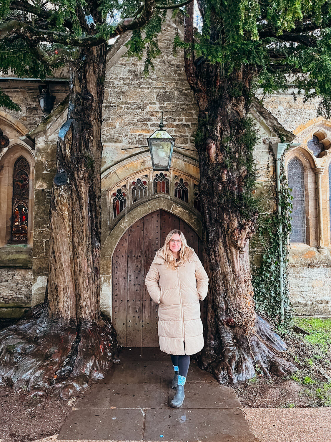 Woman in tan coat stands in front of wooden church door flanked by two large yew trees