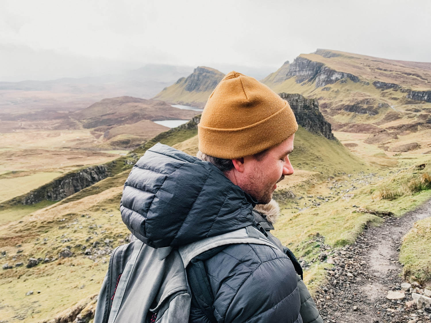 Man with black jacket and yellow hat walks on trail with green mountain in the background