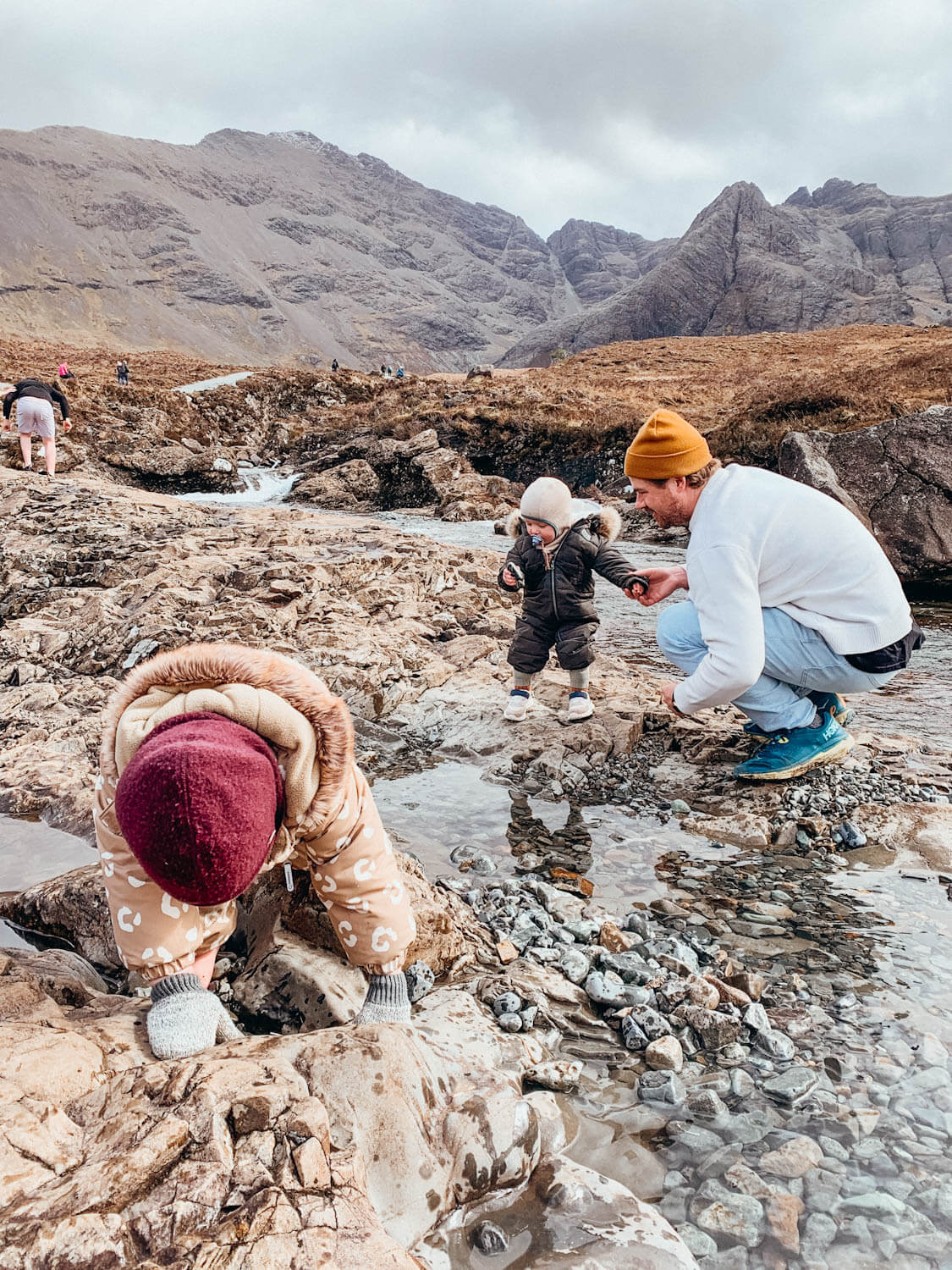 Father and two small children play in a stream with brown grass and mountains behind