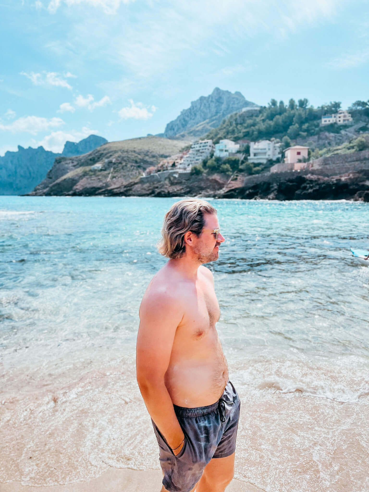 Man stands in swimsuit in front of cove with bright blue water and cliffs in the background