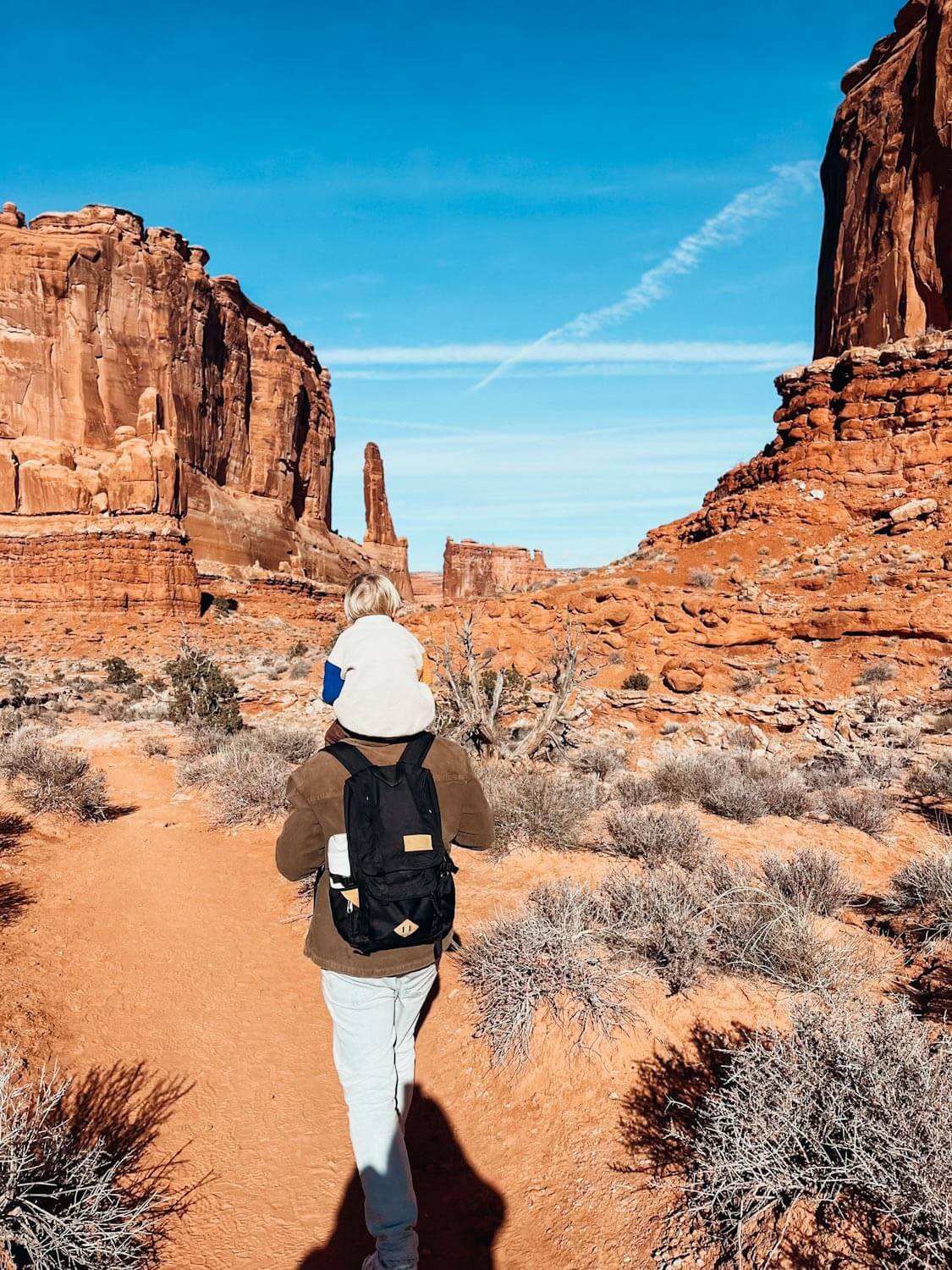 Man carries child on his shoulders on red dirt trail in Arches National Park