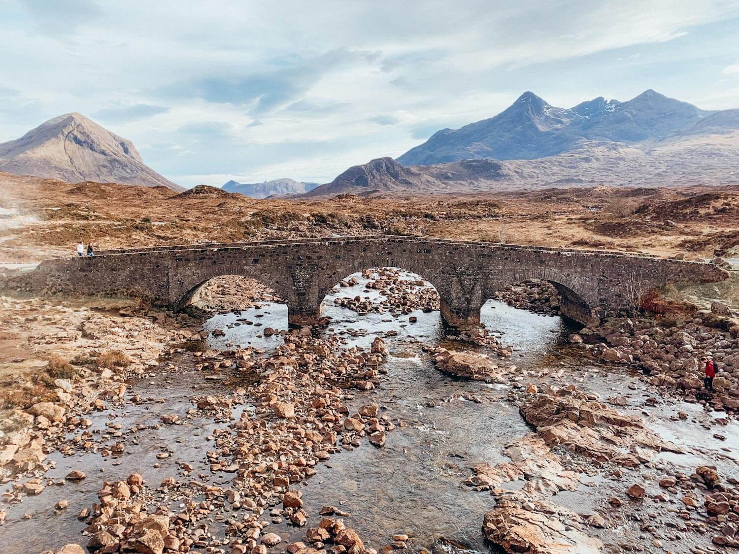Old stone bridge with three arches over a stream with brown mountains in the background