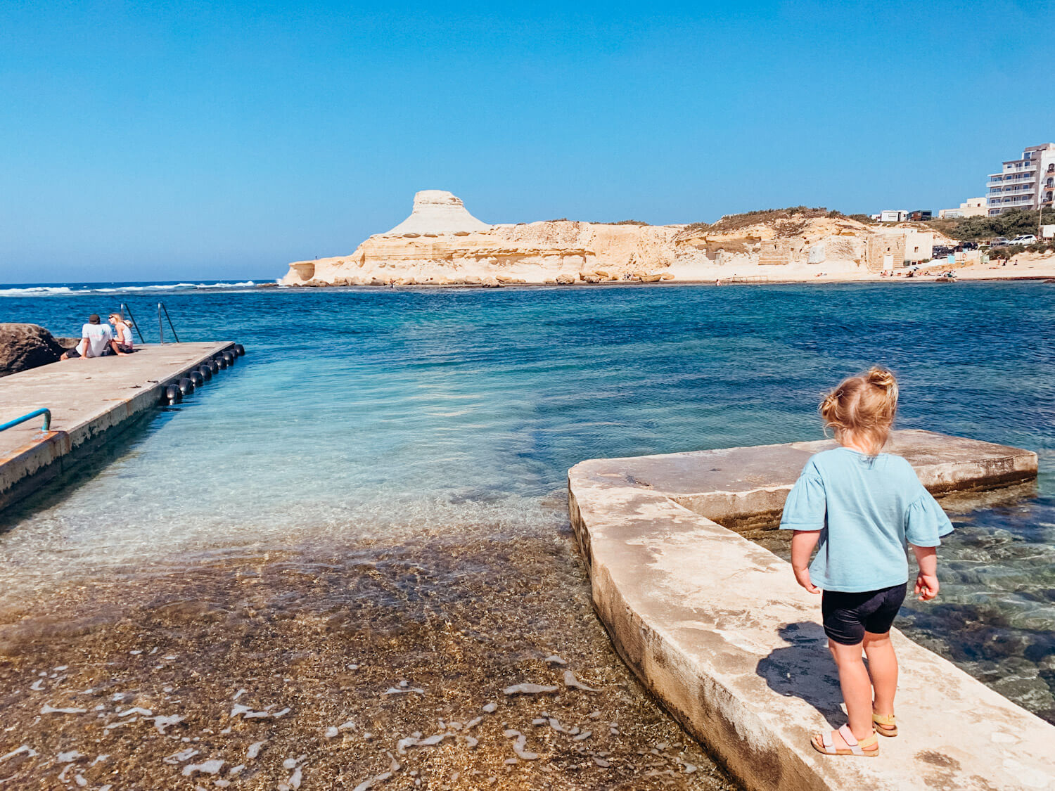 Young girl stands on a cement dock leading into bright blue water with a white rock formation in the distance