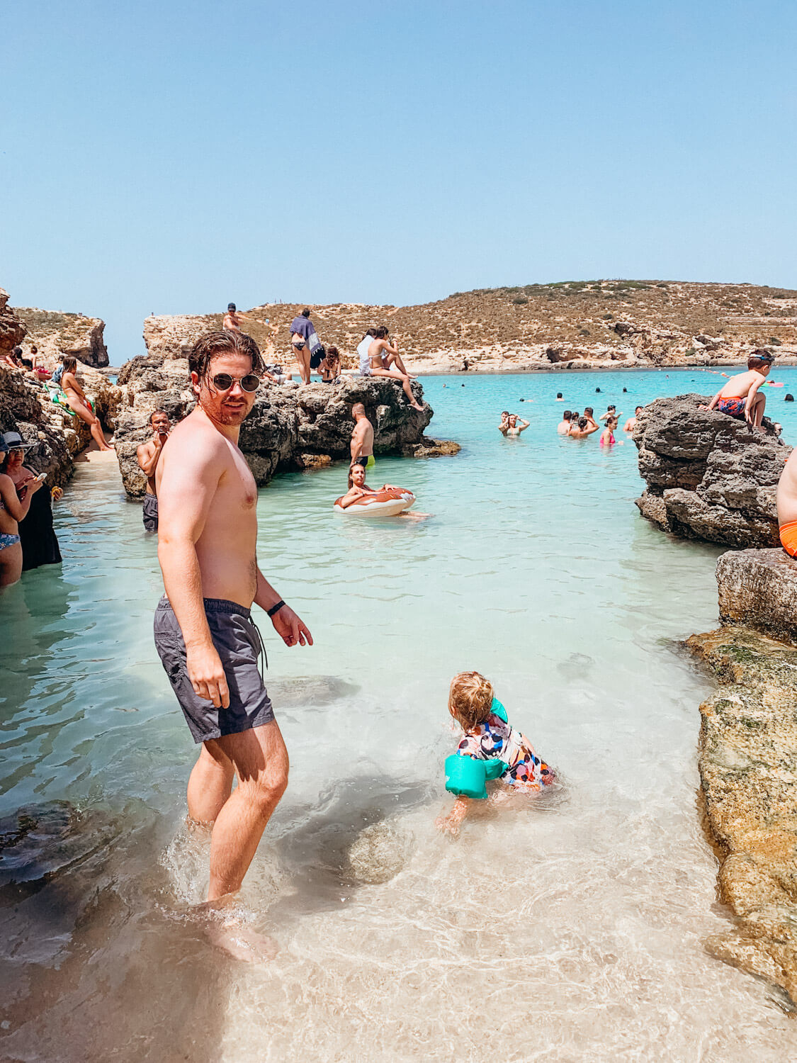 Man walks in to clear water with rocks and many people in the water