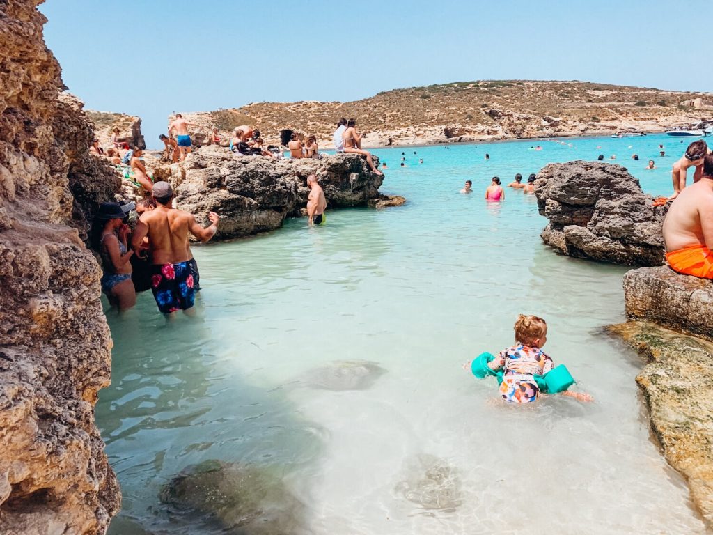 People swim in blue water and sit on rocks at the Blue Lagoon in Malta