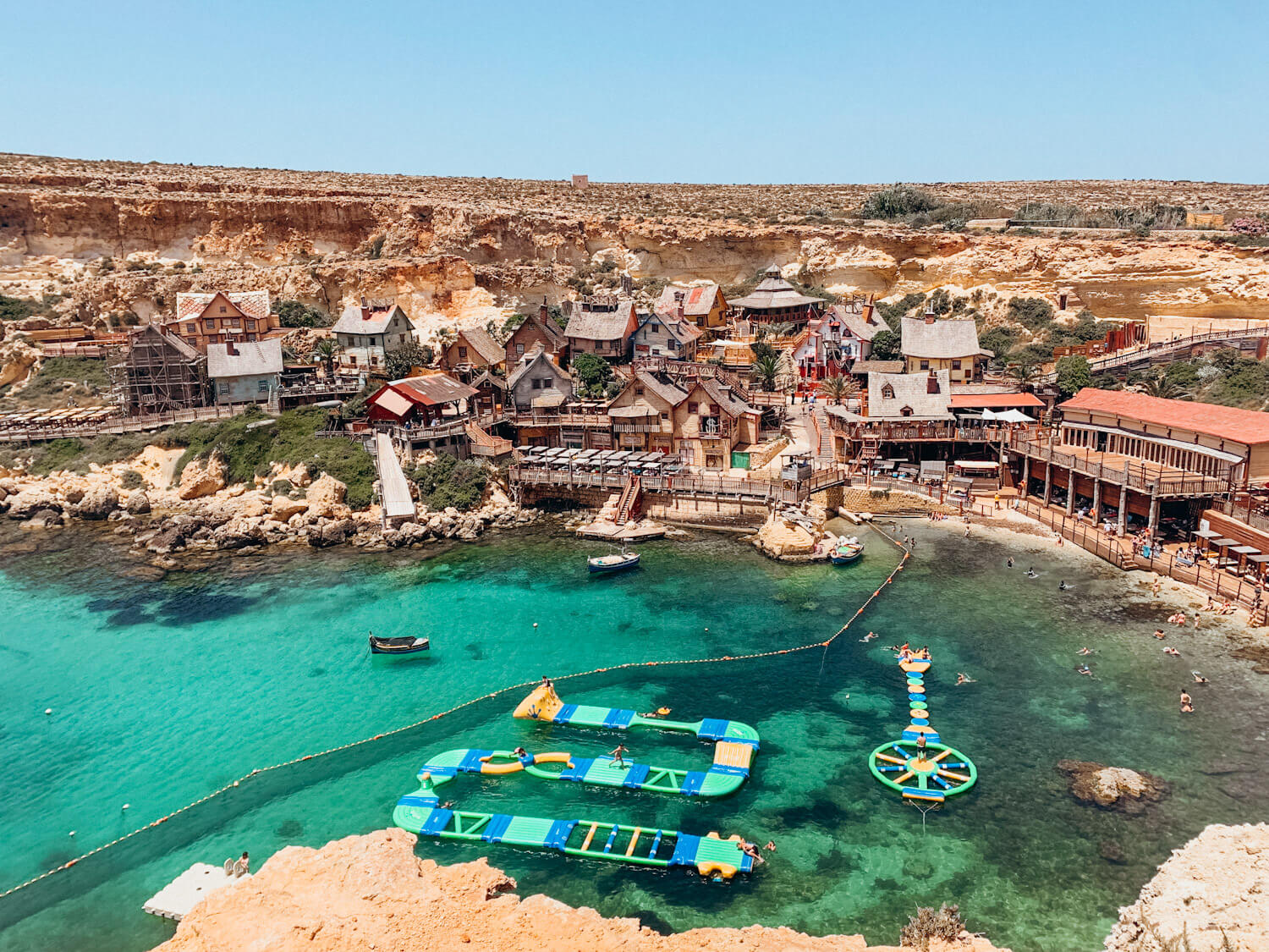 Ariel view of cove lined with wooden buildings and green water with colorful floating play spaces